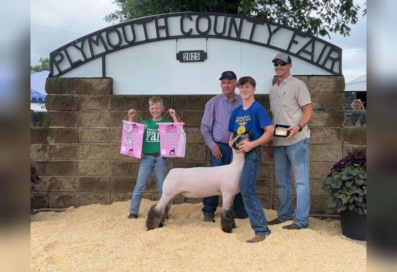 Cooper stands under the Plymouth County Fair sign, holding the head of his sheep. Also in the photo are another boy and two men.