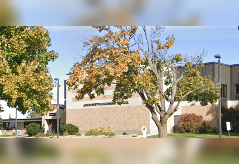 The A-W Community School is in the background, a brown and white brick building. In the foreground, partially obscuring the view of the building are trees.