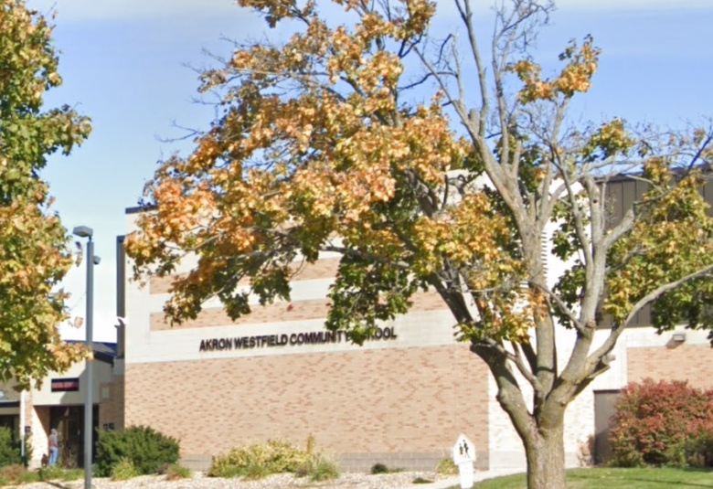 The A-W Community School is in the background, a brown and white brick building. In the foreground, partially obscuring the view of the building are trees.