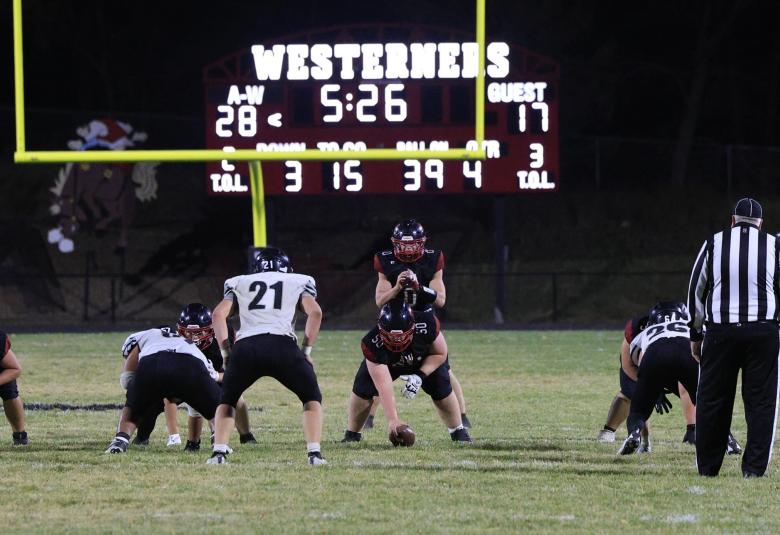 Two referees and a dozen football players are on the field, mid-game. The digital scoreboard reads "A-W 28" and "Guest 17."