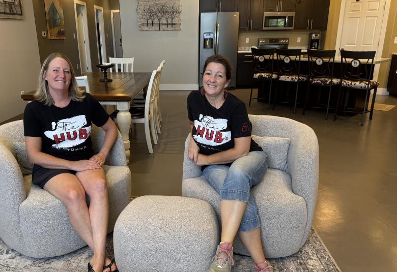 Stephanie Cacioppo and Jennifer Hart sit on stuffed chairs in the Airbnb living room. Both are wearing T-shirts that read "The HUBnb."
