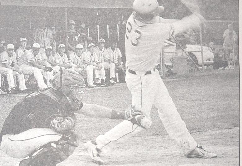 A black and white photo of a batter swinging, while the catcher stands behind them.