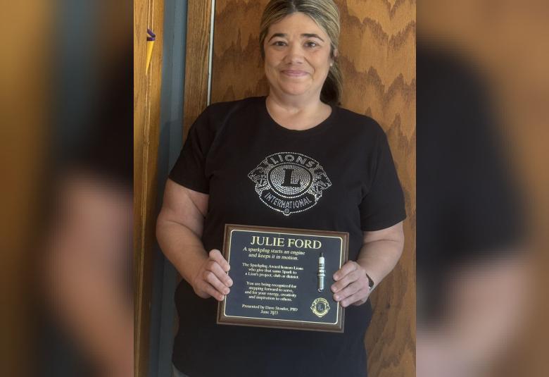 Ford stands against a wooden wall, facing the camera and holding her plaque.