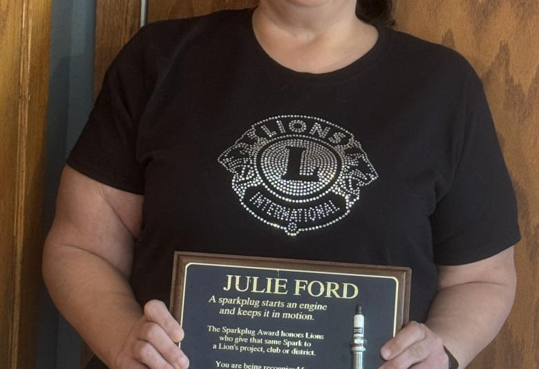 Ford stands against a wooden wall, facing the camera and holding her plaque.