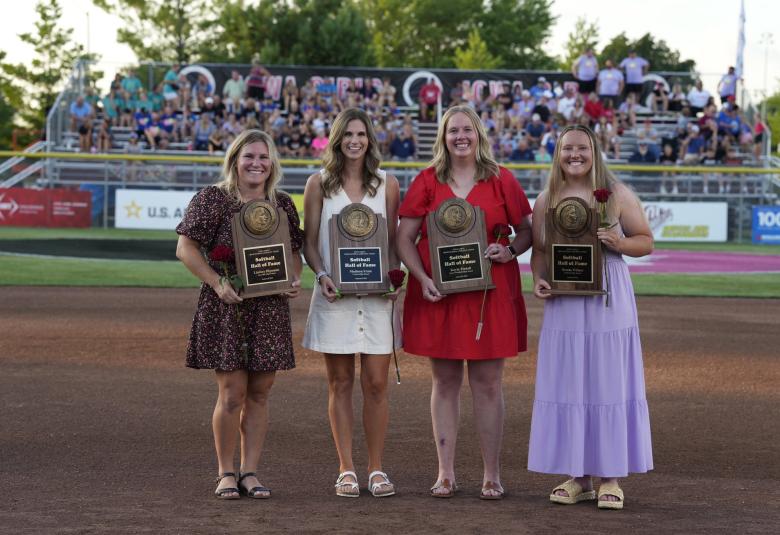 Four women stand outdoors, smiling and facing the camera. Each is holding a plaque that says "Softball Hall of Fame" and holding a red rose. In the background are spectators on bleachers.