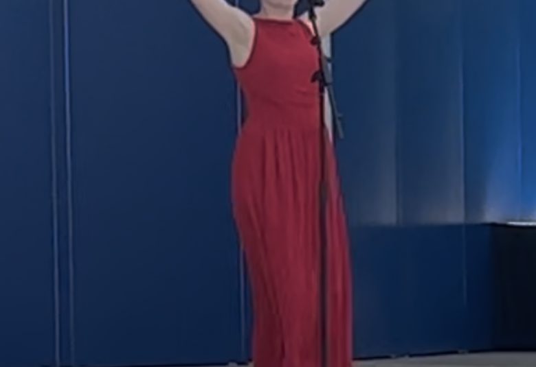 Kelsey stands on stage, wearing a red dress and singing into a microphone.