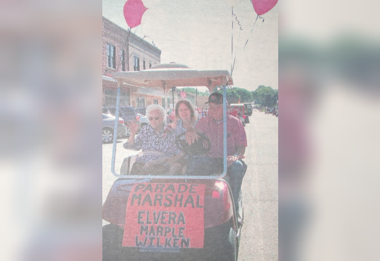 Elvera sits in a red golf cart, waving. To the right is her son, driving. Behind them in the back seat, is her daughter. There are pink balloons tied to the cart. In the front of the cart is a sign that says "Parade Marshal, Elvera Marple Wilken"