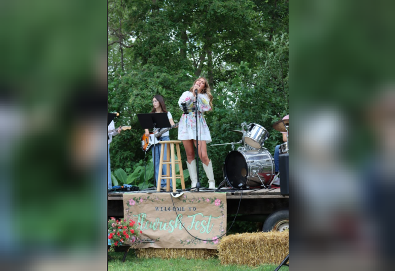 Higman stands on stage, signing into a microphone. She is wearing a white dress with a purple, pink, blue, and green floral pattern, and white cowboy boots. Hanging off the stage beneath her feet is a sign that reads "Welcome to Fleurish Fest." Behind Higman, to the left, is a woman play an electric guitar. To the left is the drum set. In the immediate background are trees, flush with green leaves.