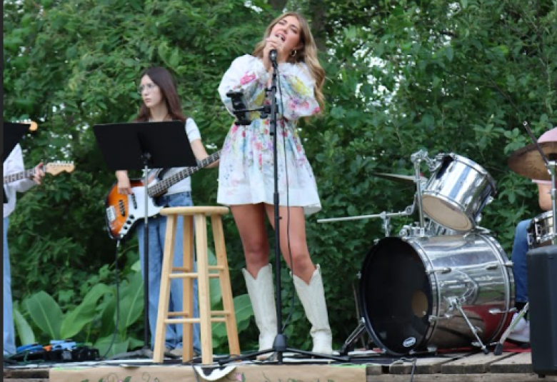 Higman stands on stage, signing into a microphone. She is wearing a white dress with a purple, pink, blue, and green floral pattern, and white cowboy boots. Hanging off the stage beneath her feet is a sign that reads "Welcome to Fleurish Fest." Behind Higman, to the left, is a woman play an electric guitar. To the left is the drum set. In the immediate background are trees, flush with green leaves.
