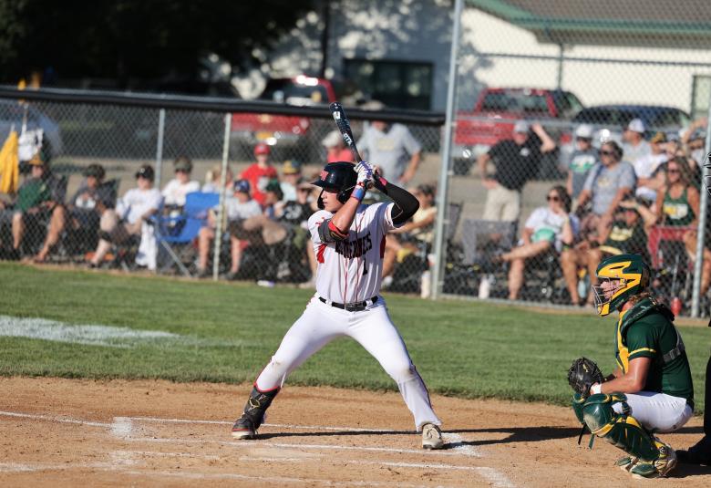 Hartman stands at bat, his body facing the camera and his turned to his right, facing the pitcher's mound. Behind him is the opposing team's catcher, who is crouched. In the background, spectators sit on lawn chairs behind a chain link fence.